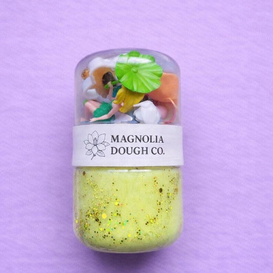 Jar of green dough with toy figures on a purple background, featuring Magnolia Dough Co. branding.