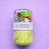 Jar of green dough with toy figures on a purple background, featuring Magnolia Dough Co. branding.