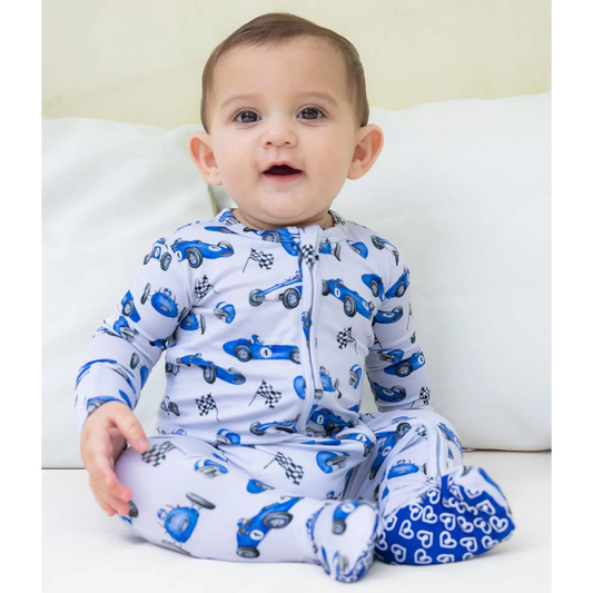 Baby wearing a footie with blue car patterns sitting on a white couch.