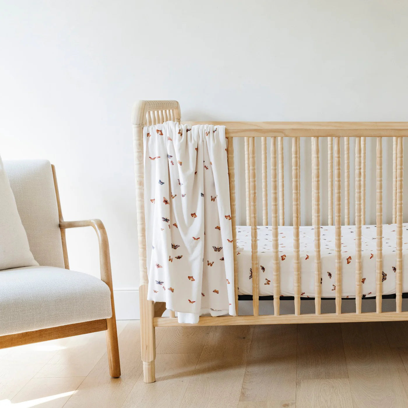 Wooden crib with a white crib sheet featuring brown patterns, next to a beige chair in a minimalistic room.