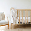 Wooden crib with a white crib sheet featuring brown patterns, next to a beige chair in a minimalistic room.