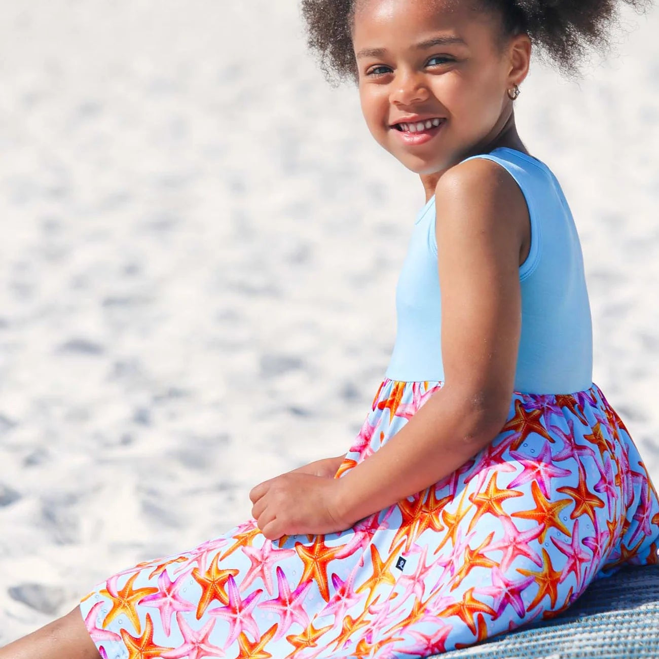 Young girl sitting on a beach wearing a light blue sleeveless top and colorful starfish-patterned shorts.